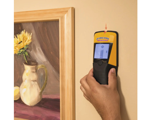 Eine Hand hält einen Zircon Ständerdetektor an einer Wand neben einem gerahmten Gemälde.