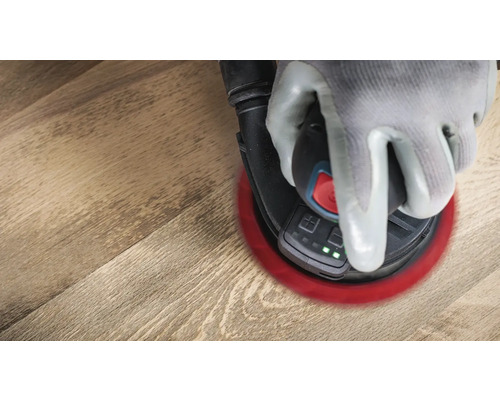 Une main gantée actionne une ponceuse excentrique sur un plancher en bois.