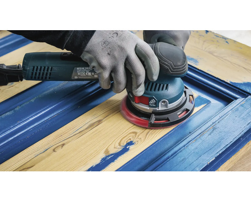 Un artisan ponce une porte en bois bleue avec une ponceuse excentrique.