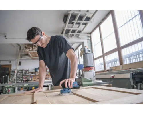 Ein Mann schleift eine Holztür mit einem elektrischen Schleifer in einer Werkstatt.