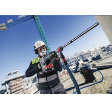 Bauarbeiter mit Atemschutz, Schutzbrille, Gehörschutz und Bauhelm bohrt mit einem Bohrhammer von Bosch auf einer Baustelle.