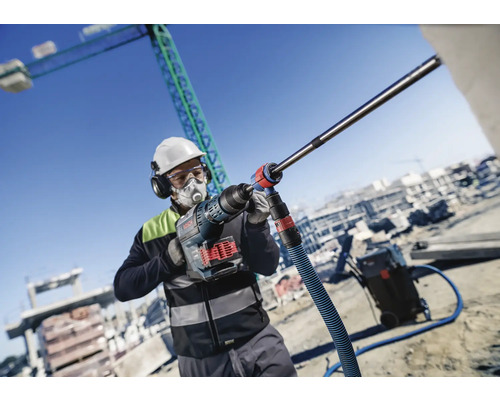 Un ouvrier du bâtiment portant un masque respiratoire et une protection auditive travaille avec une perceuse Bosch sur un chantier.