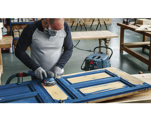Mann schleift eine blaue Tür mit einem Exzenterschleifer, Staubabsaugung ist angeschlossen.
