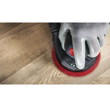 Une main gantée utilise une ponceuse excentrique sur un plancher en bois.