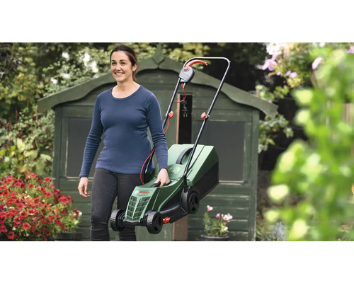 Femme portant une tondeuse à gazon électrique dans le jardin devant une remise.