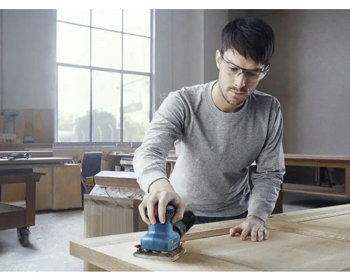 Un homme ponce du bois avec une ponceuse vibrante dans un atelier.
