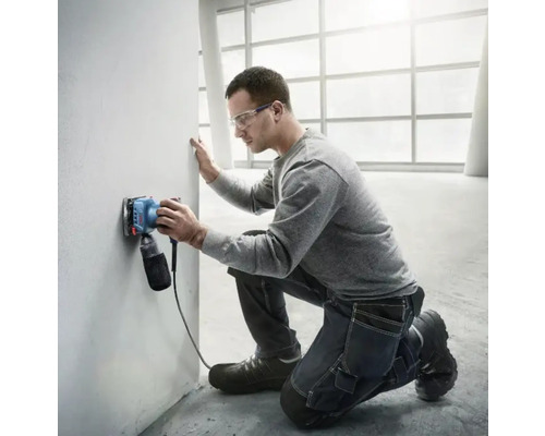 Un homme ponce un mur avec une ponceuse électrique dans une pièce lumineuse.