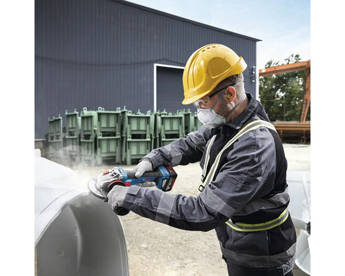 Un homme portant un casque, des lunettes de protection et un masque antipoussière meule une pièce à l'aide d'une meuleuse d'angle sans fil.