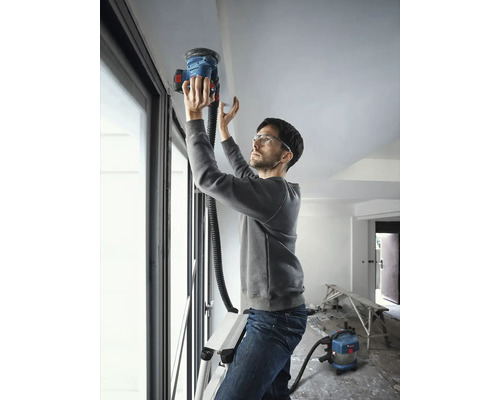 Homme sur une échelle ponçant un plafond avec une ponceuse à plafond, reliée à un aspirateur