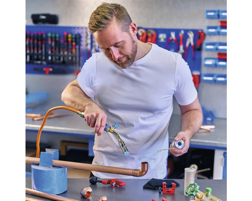 Un homme soude des tuyaux en cuivre avec un fer à souder dans un atelier, avec des raccords en cuivre et des outils sur la table.