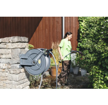 Une femme arrose des plantes avec un tuyau d''arrosage enroulé sur un dévidoir fixé à un mur de pierre dans le jardin. Logo GEKA.