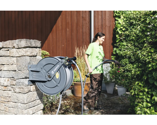 Une femme arrose des plantes avec un tuyau d''arrosage enroulé sur un dévidoir fixé à un mur de pierre dans le jardin. Logo GEKA.