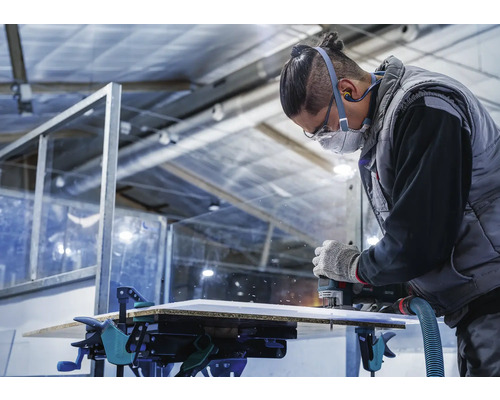 Un artisan fraise une plaque avec une défonceuse, qui est fixée avec des serre-joints sur un établi.