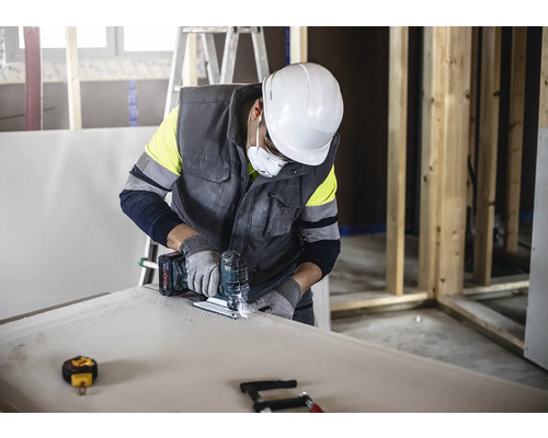 Un artisan coupe une plaque de plâtre avec une scie sauteuse tout en portant des vêtements de protection.