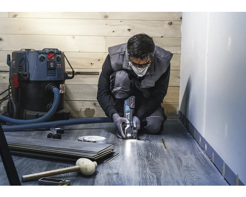 Un homme agenouillé coupe du stratifié avec un outil multifonction. Un aspirateur, des planches de stratifié et des outils sont également visibles.