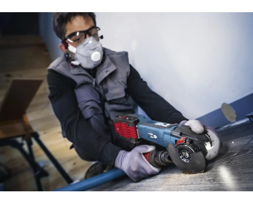 Un homme travaille avec une meuleuse d''angle sur un revêtement de sol, des équipements de protection individuelle sont présents