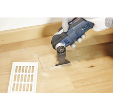 Une personne découpe un trou dans un plancher en bois à côté d''une grille de ventilation à l''aide d''un outil multifonction.