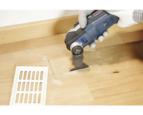 Une personne découpe un trou dans un plancher en bois à côté d''une grille de ventilation à l''aide d''un outil multifonction.