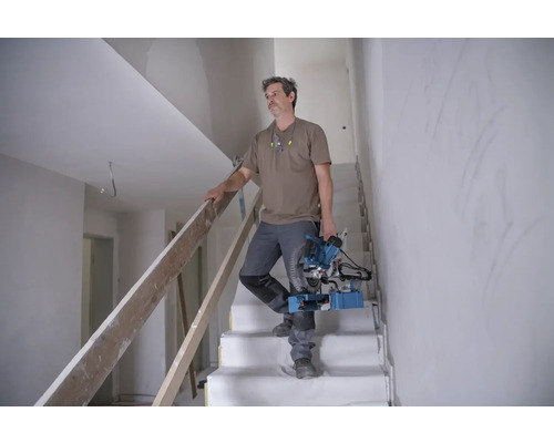 Un artisan descend un escalier avec une scie à onglets dans une maison en construction.