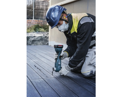 Un ouvrier du bâtiment portant un casque de sécurité et un masque facial utilise un outil multifonction pour découper une rainure dans une lame de terrasse.
