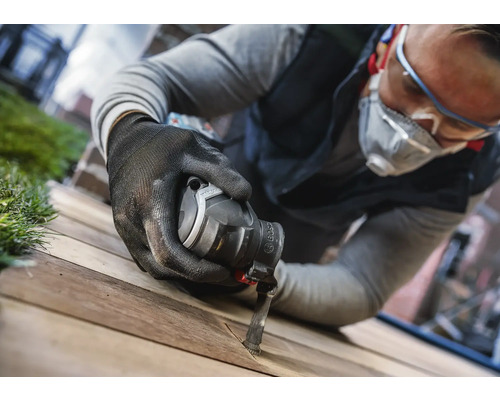 Un homme portant des lunettes de protection, un masque respiratoire et des gants travaille sur une planche de bois avec un outil multifonction.