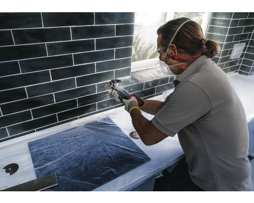 Fliesenleger bei der Arbeit mit einem Multifunktionswerkzeug an einer Wand mit schwarzen Fliesen.