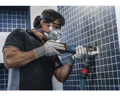 Un homme enlève des carreaux avec un outil multifonction, équipé de lunettes de protection, de protections auditives, d''un masque antipoussière et de gants