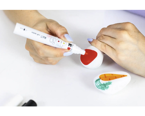 Une personne peint une pierre avec un feutre rouge, tandis qu''une autre pierre avec une carotte se trouve au premier plan.