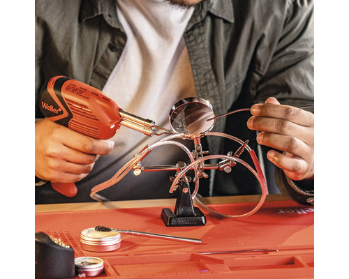 Une personne soude des fils avec un pistolet à souder, qui sont fixés avec un support équipé d''une loupe. Des flux et des accessoires de nettoyage se trouvent sur la surface de travail.