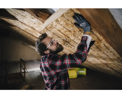 Un homme portant des lunettes de protection et des gants fixe de la laine isolante sur un toit.