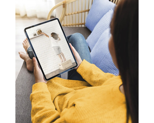 Frau betrachtet Video auf Tablet, das ein Kind zeigt, das auf einer Kiste steht, um Gegenstände von einem Regal zu erreichen