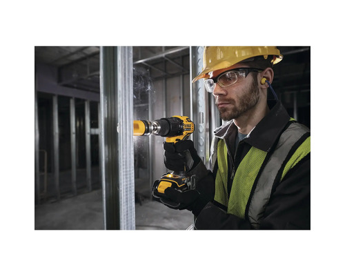 Ein Mann bohrt mit einer Bohrmaschine ein Loch in eine Metallwand. Er trägt eine Schutzbrille, einen Helm und Ohrstöpsel.