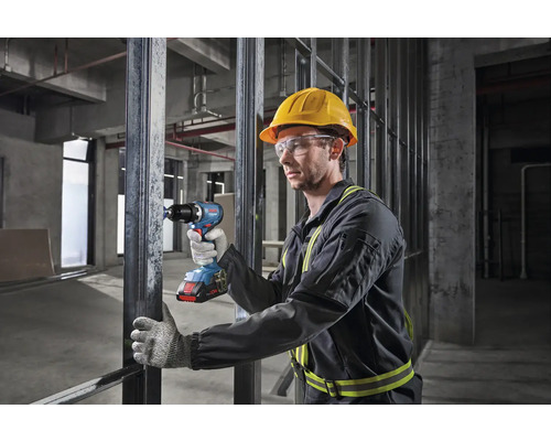 Un ouvrier portant un casque et des lunettes de sécurité utilise une visseuse sans fil sur un chantier de construction.