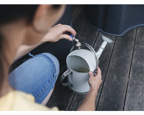 Eine Person befüllt eine weiße Gießkanne mit einem Wasserhahn neben einem Wassertank.