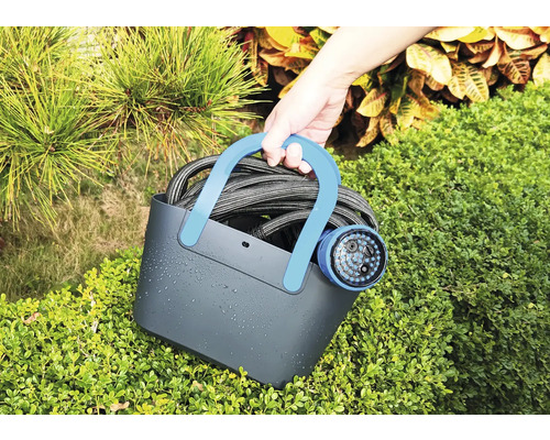 Person hält einen Wasserschlauch mit Gartenspritze in einem Tragekorb vor Büschen.