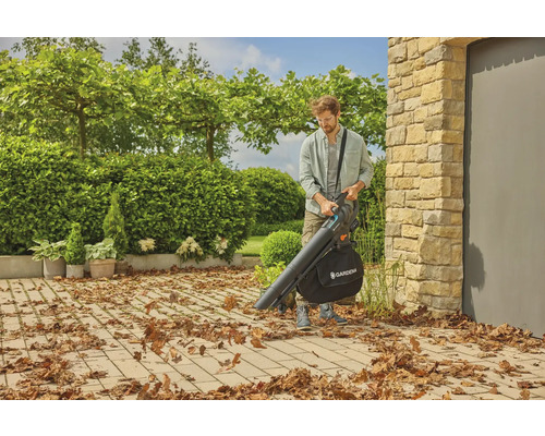 Un homme utilise un aspirateur de feuilles de la marque Gardena dans un jardin avec des arbres et des buissons.