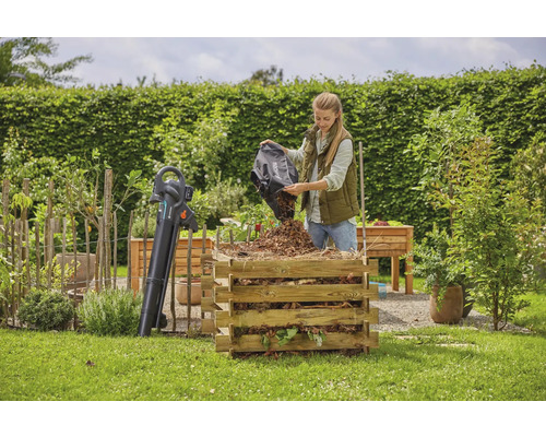 Une femme vide des feuilles dans un composteur en bois dans un jardin. Un souffleur de feuilles se trouve à côté.