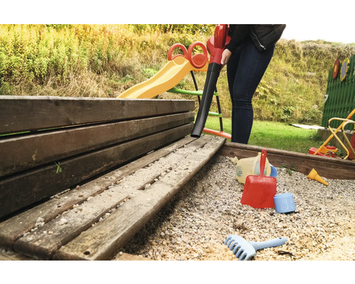 Eine Person reinigt einen Sandkasten mit einem Laubsauger in einem Garten.