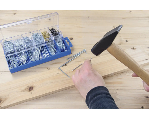 Hammer schlägt Nagel in Holzbrett neben Sortimentskasten mit Nägeln.