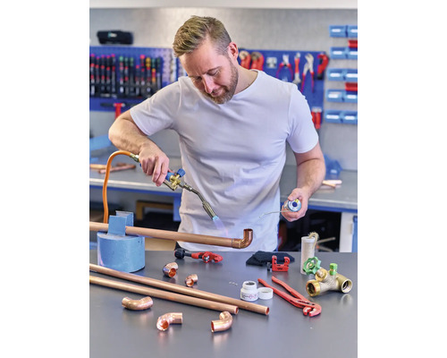 Ein Installateur lötet Kupferrohre mit einem Schweißbrenner, während verschiedene Werkzeuge und Rohrstücke auf der Werkbank liegen.