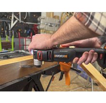 Une personne utilise un outil multifonction de marque Worx pour travailler une pièce de bois. L''outil est noir et orange.