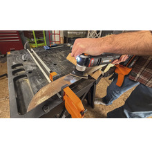 Une personne utilise un outil multifonction pour poncer une pièce fixée par des pinces sur un établi.
