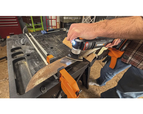 Une personne utilise un outil multifonction pour poncer une pièce fixée par des pinces sur un établi.