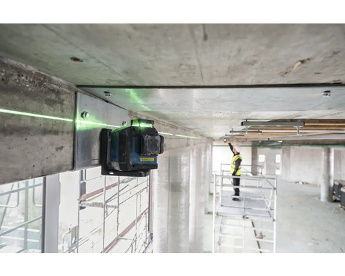 Ein grüner Linienlaser ist an einem Betonbalken befestigt und projiziert eine grüne Laserlinie. Im Hintergrund steht ein Arbeiter auf einem Gerüst.