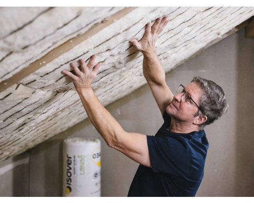 Mann befestigt Dämmwolle an einer Dachschräge. Eine Rolle Dämmmaterial ist im Hintergrund sichtbar.