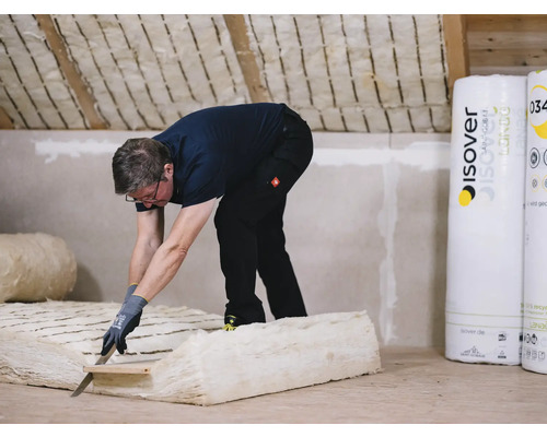 Ein Mann schneidet Dämmwolle mit einem Messer in einem Dachgeschoss. Im Hintergrund sind Rollen von Dämmmaterial der Marke Isover zu sehen.