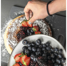 Eine Hand dekoriert einen Kuchen mit Puderzucker, Erdbeeren, Blaubeeren und Brombeeren.