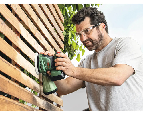 Un homme ponce une surface en bois à l'extérieur avec une ponceuse excentrique