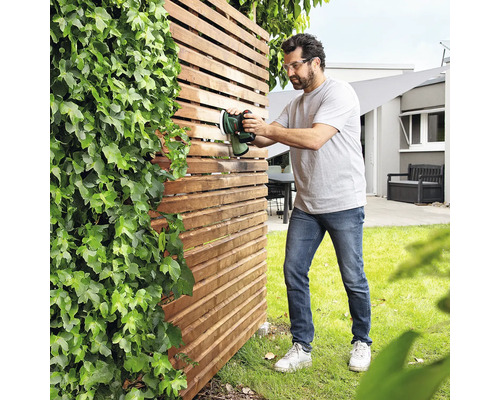 Un homme traite une clôture en bois avec un pulvérisateur de peinture dans le jardin.