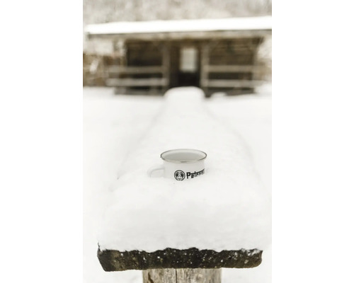 Tasse en émail avec le logo Petronam dans la neige devant une cabane en bois